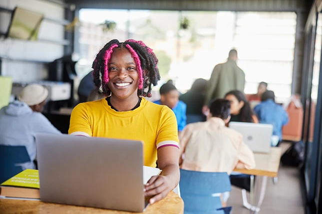 Portrait of a young female college student with braided hair smiling while doing her homework on a laptop in a busy campus cafeteria